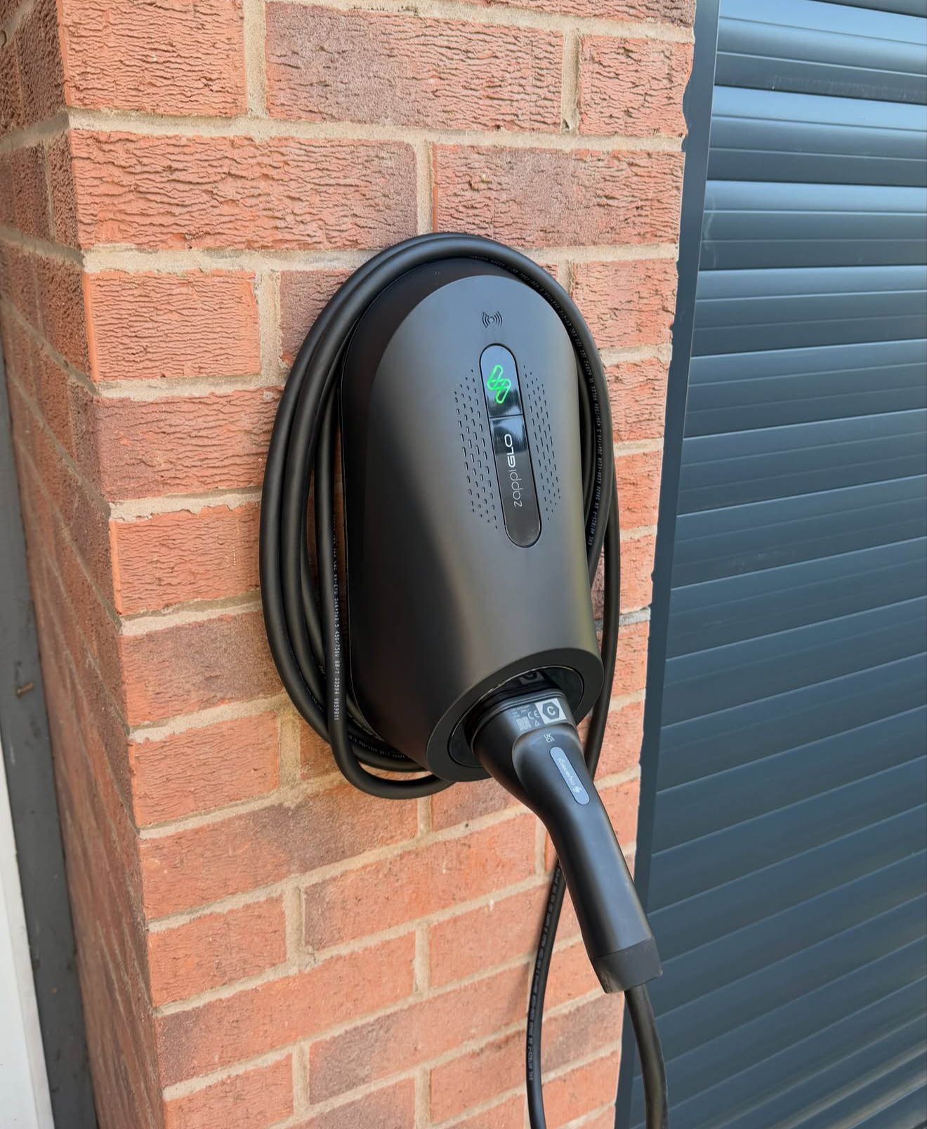 Zappi GLO EV charger mounted on a red brick wall beside a garage door